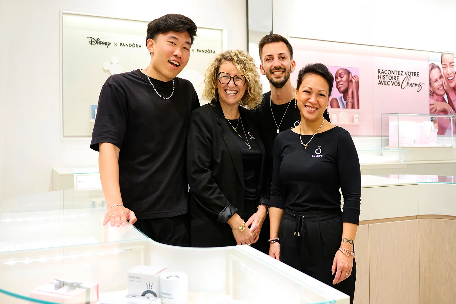 Four smiling staff members in a Disney x Pandora store stand behind a glass display case showing Pandora jewelry.