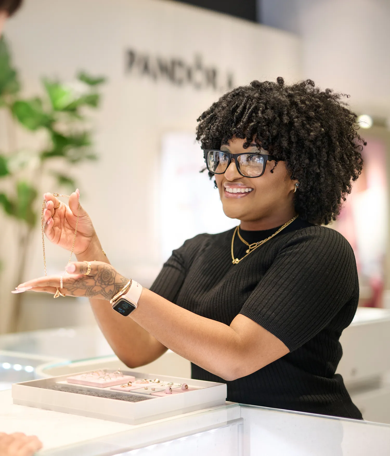 A woman with curly hair and glasses holds up a gold necklace for a customer in a jewelry store