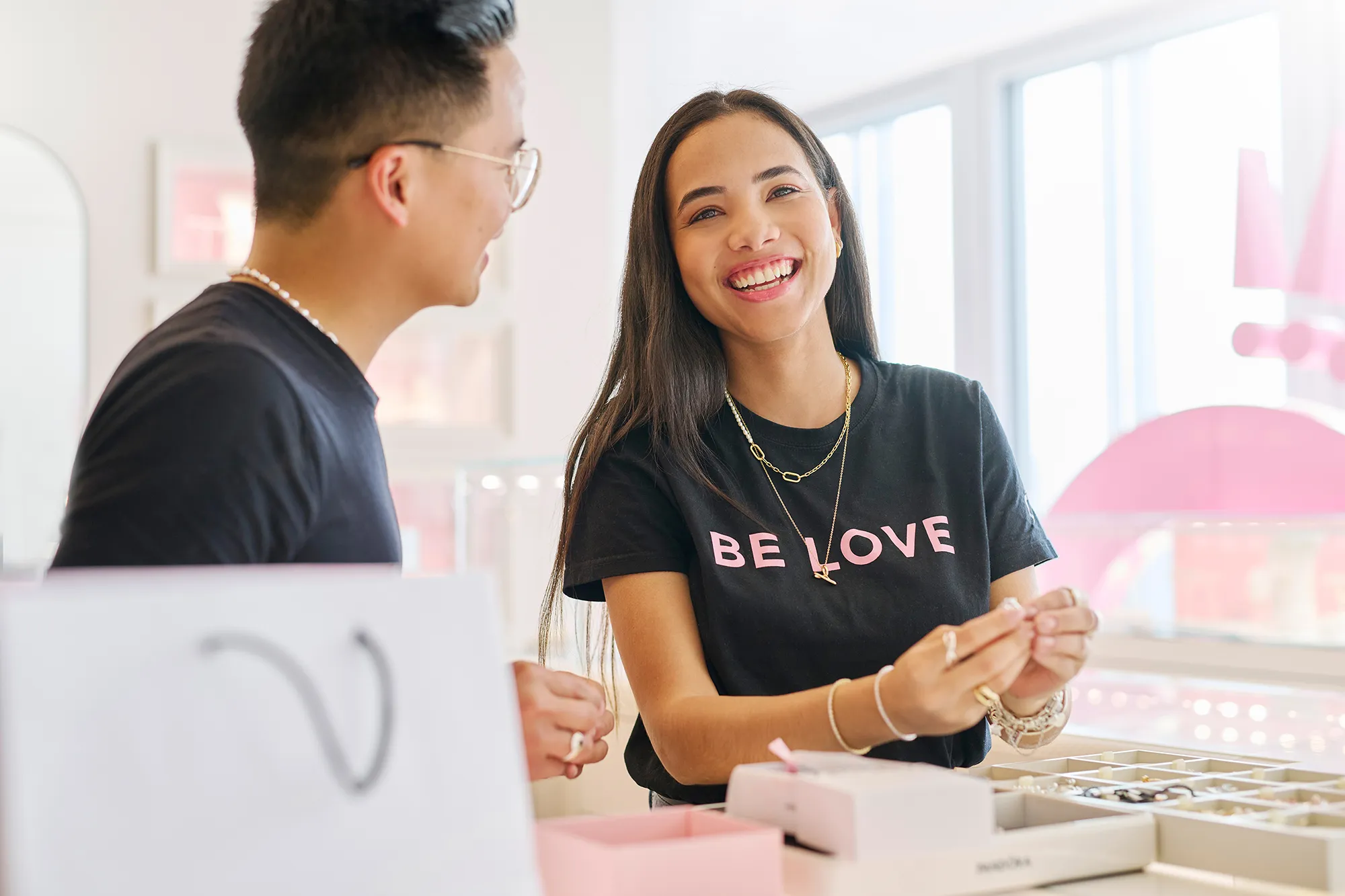A smiling woman wearing a black t-shirt with "BE LOVE" printed in pink texts is holding a ring in her hands.