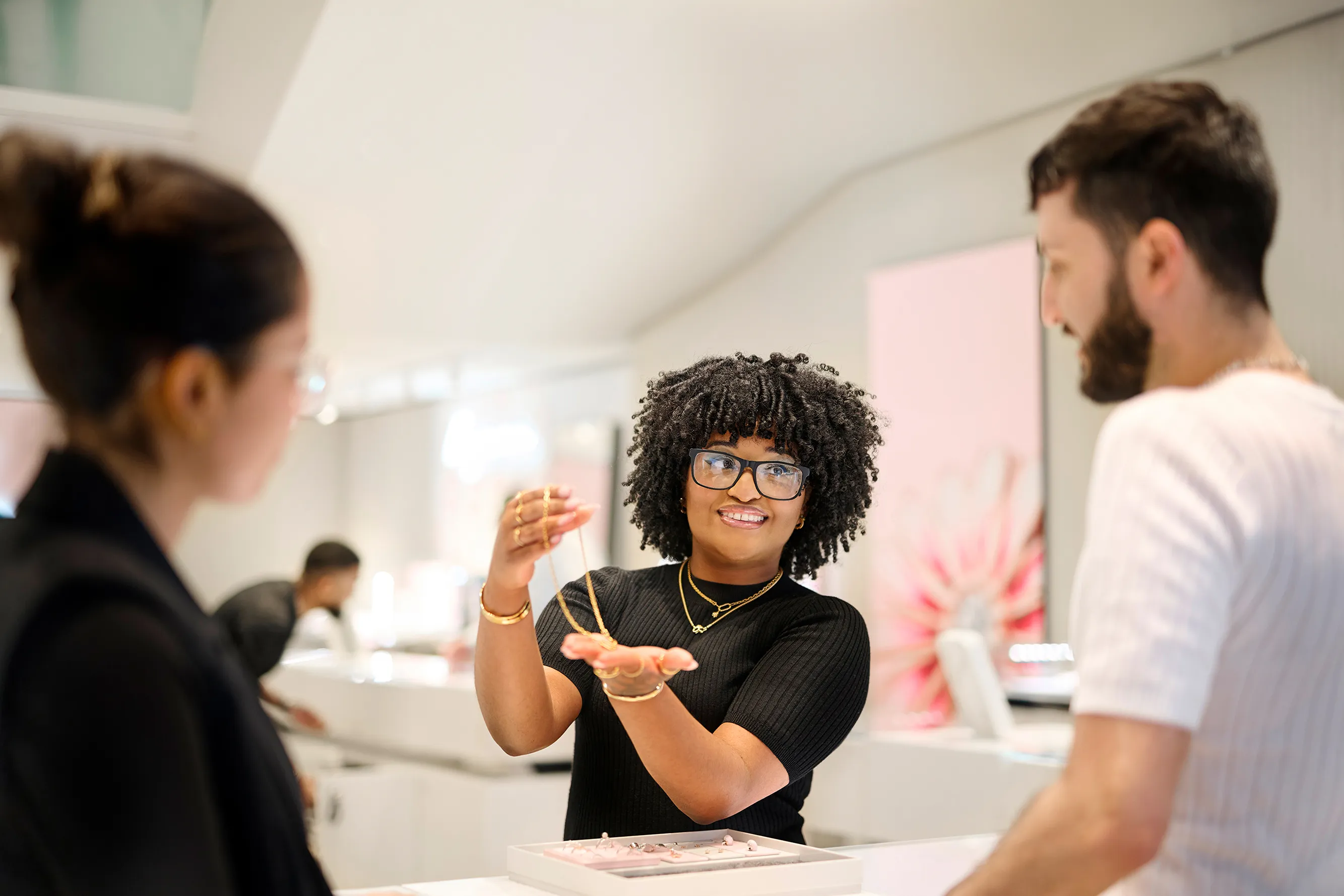 A smiling sales associate shows two women a display of jewelry, including necklaces and rings.