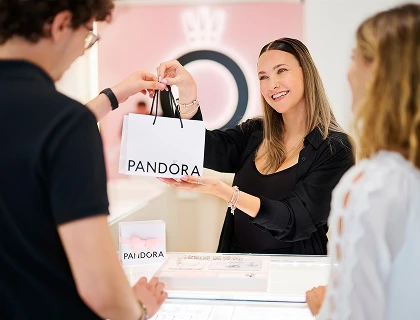A smiling Pandora employee hands a shopping bag to a customer at the jewelry store counter