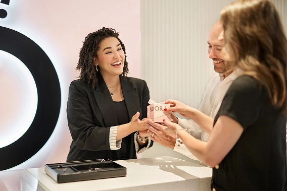 A smiling woman wearing a black t-shirt with "BE LOVE" printed in pink and a man wearing a black t-shirt stand in a jewelry store.