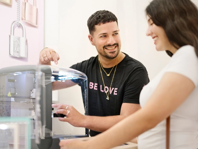 A man in a black t-shirt with the words "BE LOVE" points at a 3D printer while a smiling woman in a white top looks on
